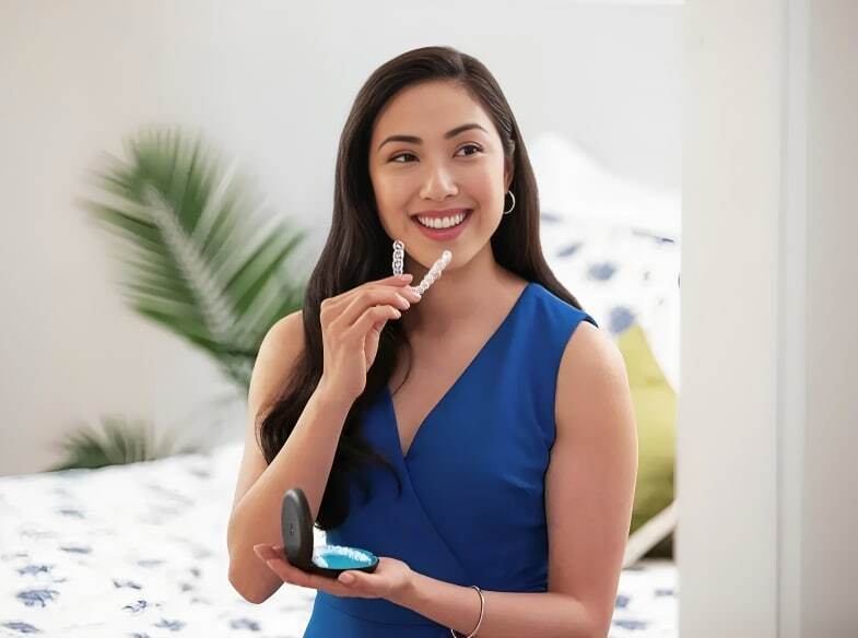 Woman holding Invisalign clear aligner tray while smiling in modern dental office.