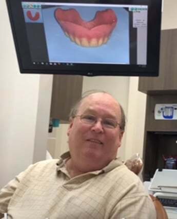 Male dentist smiling in clinical office with digital smile design displayed on monitor behind him.