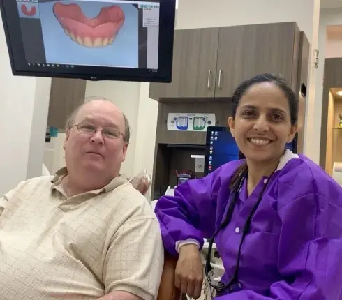 Dentist and patient smiling in dental treatment room with digital smile design displayed on monitor.