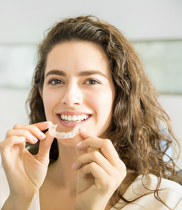 Young woman holding clear Invisalign aligner and smiling at camera in dental office.